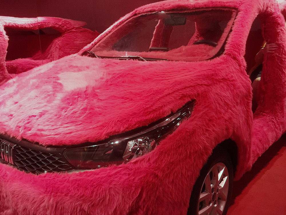Automóvel inteiramente coberto por pelúcia rosa em exposição na mostra comemorativa dos 50 anos da Fiat no Brasil. A fotografia destaca a parte frontal do veículo, evidenciando o contraste entre a textura felpuda da instalação artística e os faróis, grade e rodas originais do carro.