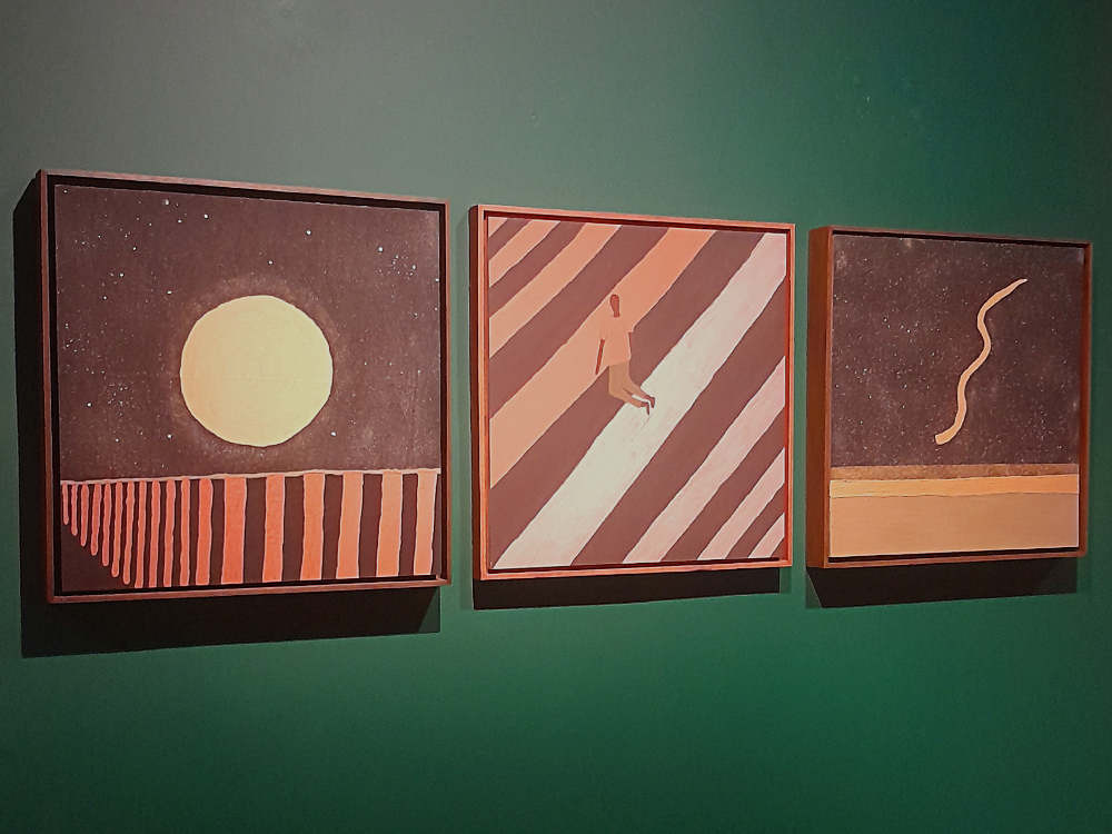 Conjunto de três pinturas geométricas expostas lado a lado sobre uma parede verde-escura. A primeira apresenta um grande círculo claro semelhante ao sol ou à lua sobre um céu estrelado; a segunda mostra uma figura humana caminhando entre faixas diagonais; e a terceira retrata uma linha sinuosa sobre uma paisagem minimalista. As obras exploram formas simples, cores terrosas e composições abstratas.