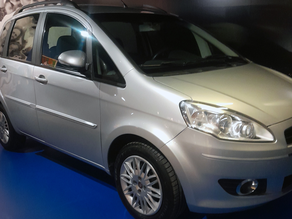 Fiat Idea prata exposto na exposição comemorativa dos 50 anos da Fiat no Brasil. O veículo está estacionado sobre piso azul, iluminado por luzes do museu, destacando o design característico da minivan e sua importância na história da marca. Ao fundo, o ambiente escuro direciona a atenção para o automóvel.