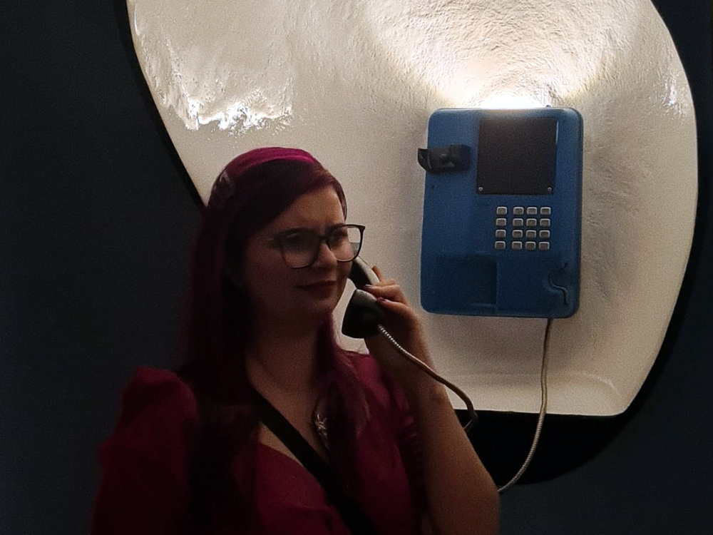 Mulher de cabelos rosa escuro, usando óculos e macacão rosa, segura o fone de um orelhão azul instalado como obra interativa da exposição. Atrás dela há um grande elemento branco em formato orgânico iluminado, criando contraste com o ambiente escuro da galeria. A fotografia transmite um momento divertido de interação com a instalação.