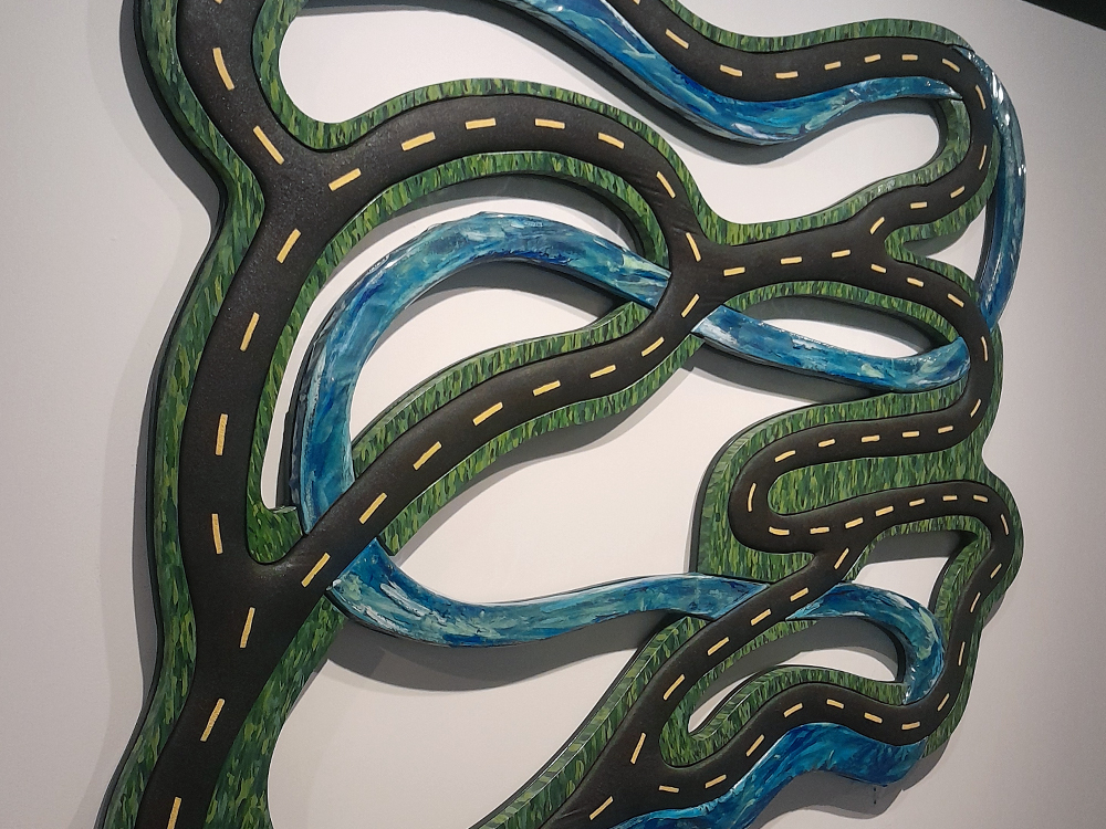 Obra de arte em relevo representa uma malha de estradas sinuosas cruzando rios azuis sobre áreas verdes, formando um desenho orgânico semelhante a um mapa visto de cima. As vias possuem marcações amarelas centrais e serpenteiam por toda a composição, evocando viagens de carro, território de brasilidade e paisagens naturais.