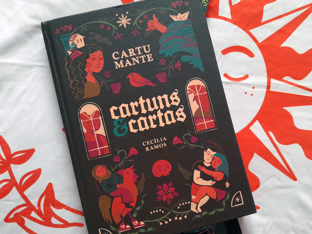 Capa do livro Cartumante: Cartuns & Cartas, de Cecília Ramos, exibindo ilustrações coloridas de figuras humanas, pássaros, flores e elementos gráficos sobre fundo escuro. O livro está apoiado em um tecido vermelho com estampa de sol, destacando o contraste entre as cores vibrantes da arte e o fundo.
