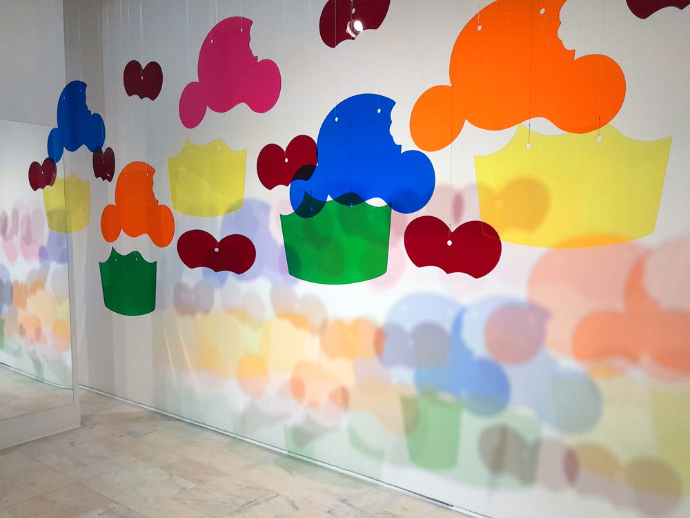 Instalação com peças coloridas em formato de Bolinho penduradas por fios transparentes, formando um conjunto de elementos acrílicos em tons de vermelho, rosa, laranja, amarelo, verde e azul. As sombras projetadas na parede branca criam camadas de cor e profundidade no espaço expositivo.