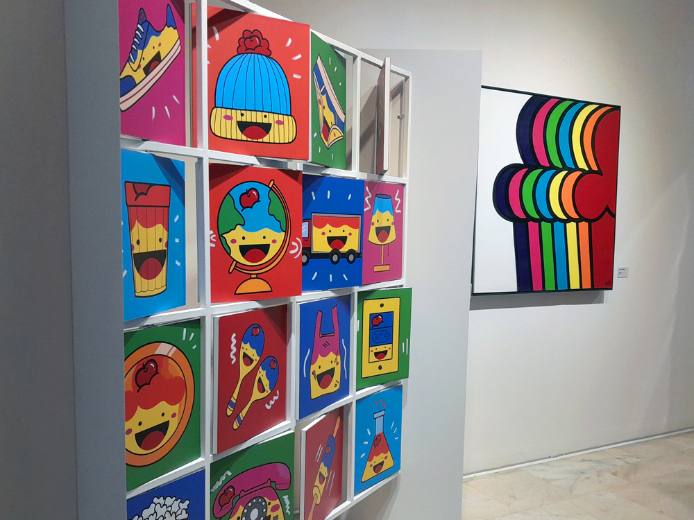 Parede com dezesseis quadros pop?art exibindo objetos do cotidiano em forma do personagem com rostos sorridentes, como sapato, gorro, lápis, globo, caminhão e telefone, cada um sobre fundo colorido, formando um jogo da memporia. Ao lado, obra abstrata formada por formas sobrepostas em tons de arco?íris, compondo uma área vibrante da exposição.