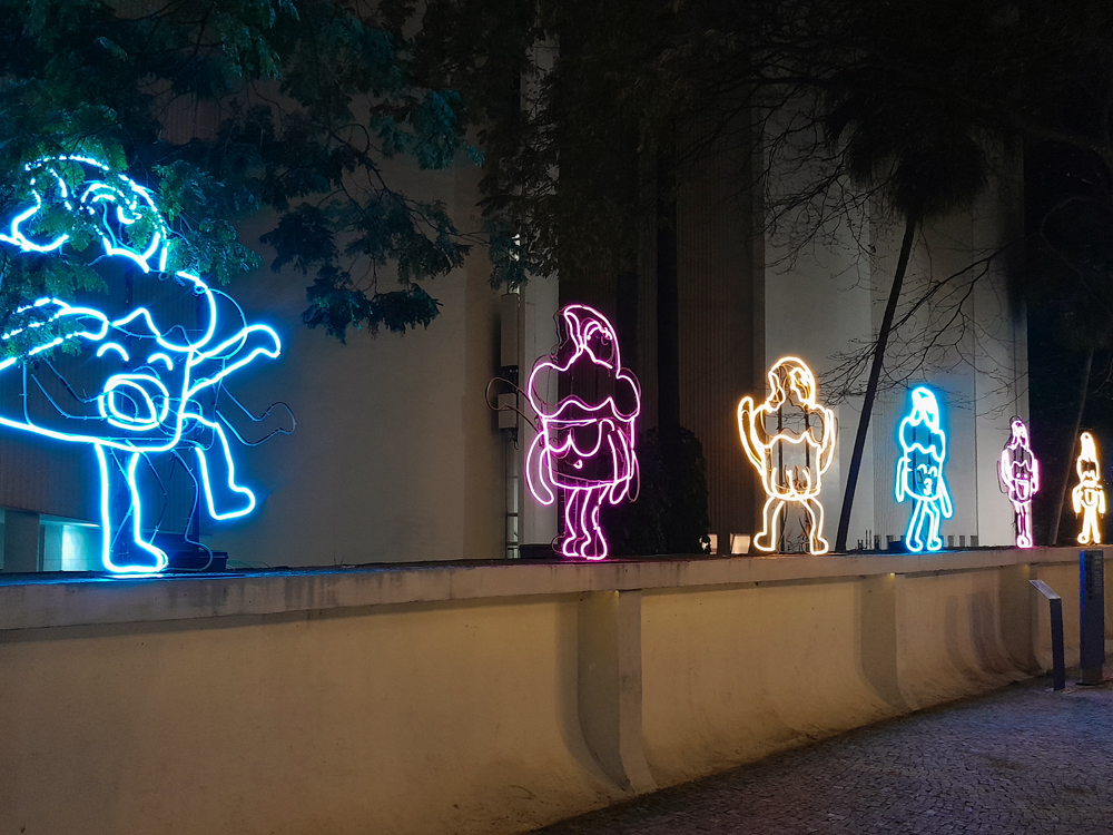Instalação noturna com figuras do personagem estilizadas feitas em luz neon nas cores azul, rosa, amarelo e laranja, alinhadas sobre um muro baixo. Árvores e prédio com janelas altas aparecem ao fundo, destacando o brilho das luzes no ambiente externo.