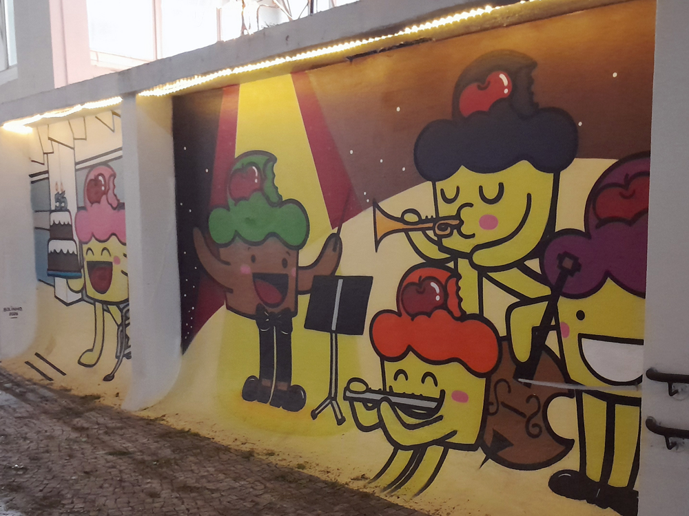 Mural colorido do Bolinho em formato de cupcake realizando atividades musicais, como cantar, reger e tocar instrumentos como trompete, flauta e violoncelo. O fundo em amarelo e vermelho cria efeito de palco iluminado, enquanto à esquerda outro Bolinho aparece em cenário de cozinha.