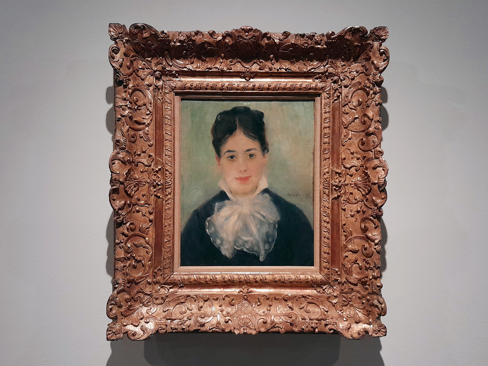 Retrato emoldurado de pessoa com cabelo escuro preso e roupa escura com grande laço branco no pescoço. O fundo é verde suave, e a moldura apresenta entalhes elaborados em estilo clássico.