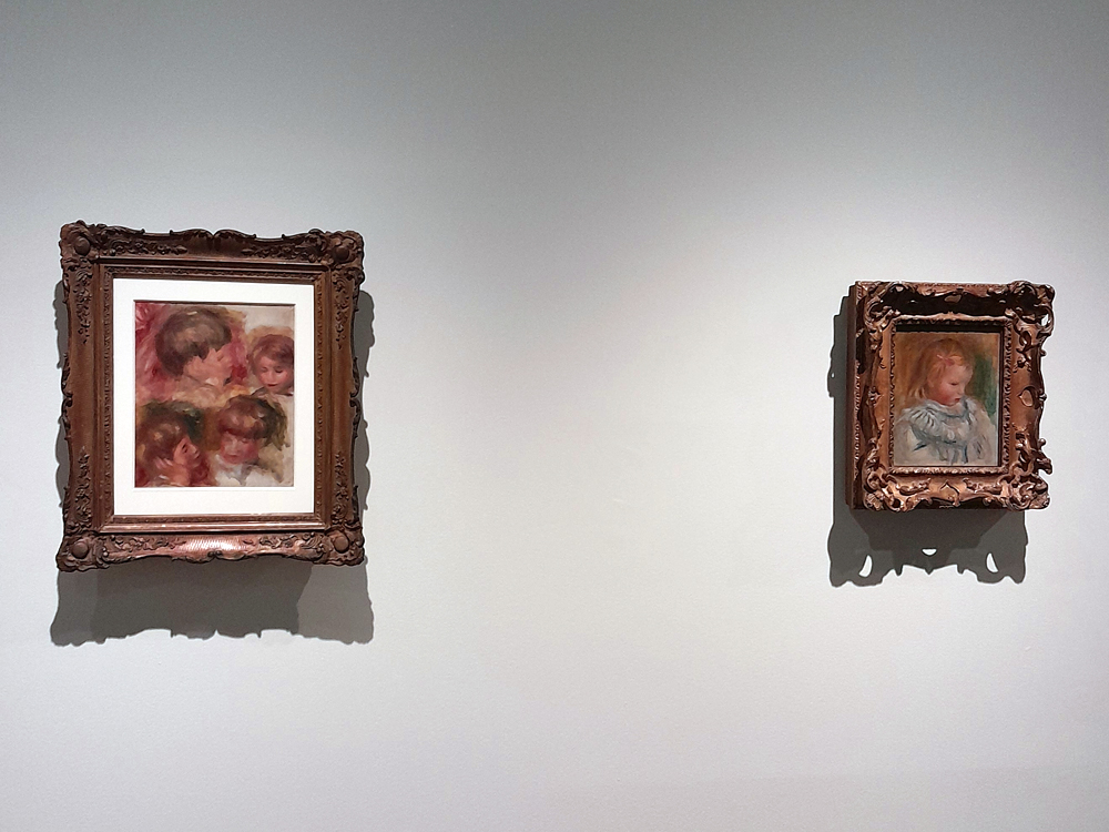 Dois quadros impressionistas de Renoir em molduras ornamentadas expostos em parede clara: à esquerda, composição com várias crianças; à direita, retrato de criança de cabelos claros usando roupa azul, ambos com pinceladas suaves e cores delicadas.