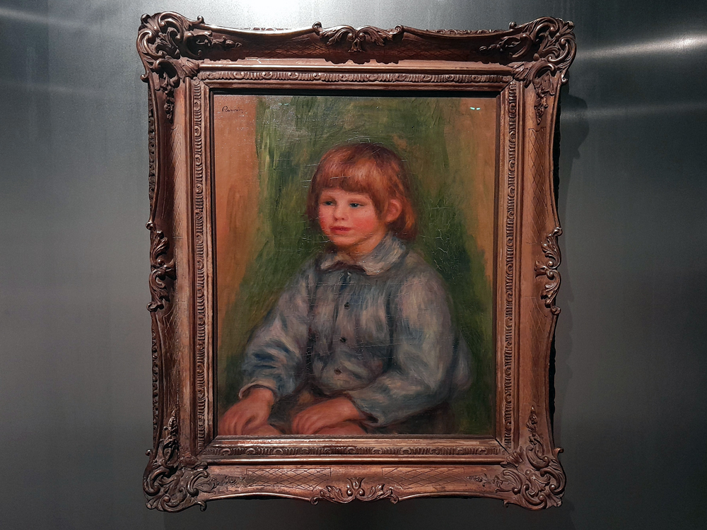 Pintura emoldurada de criança com cabelos claros e camisa azul?acinzentada, sentada com as mãos apoiadas à frente. O fundo mistura tons verdes e marrons, e a moldura apresenta entalhes florais ornamentados.