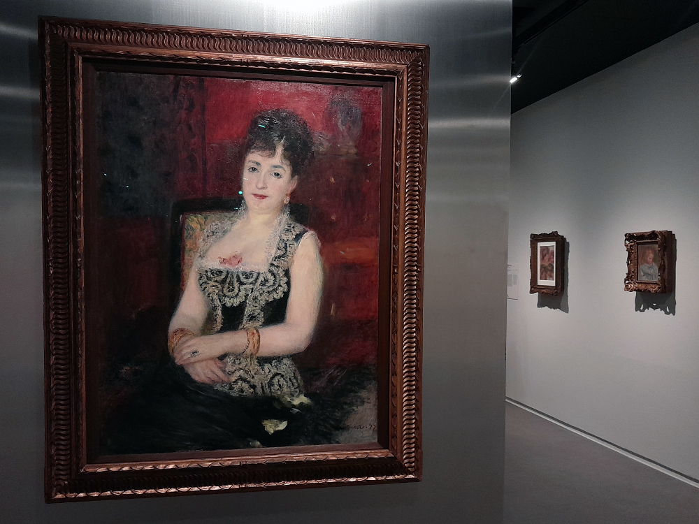 Interior de galeria da exposição Renoir com pintura em destaque mostrando figura sentada usando vestido preto com bordados dourados sobre fundo vermelho. Ao fundo, duas pinturas menores em molduras ornamentadas completam a parede expositiva.