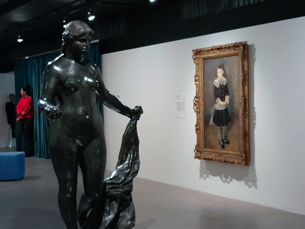 Sala de galeria com escultura em bronze de figura nua segurando tecido ao lado de pintura emoldurada de jovem em traje escuro com detalhes de renda. A parede branca, cortinas azuis e visitantes ao fundo compõem o ambiente expositivo.