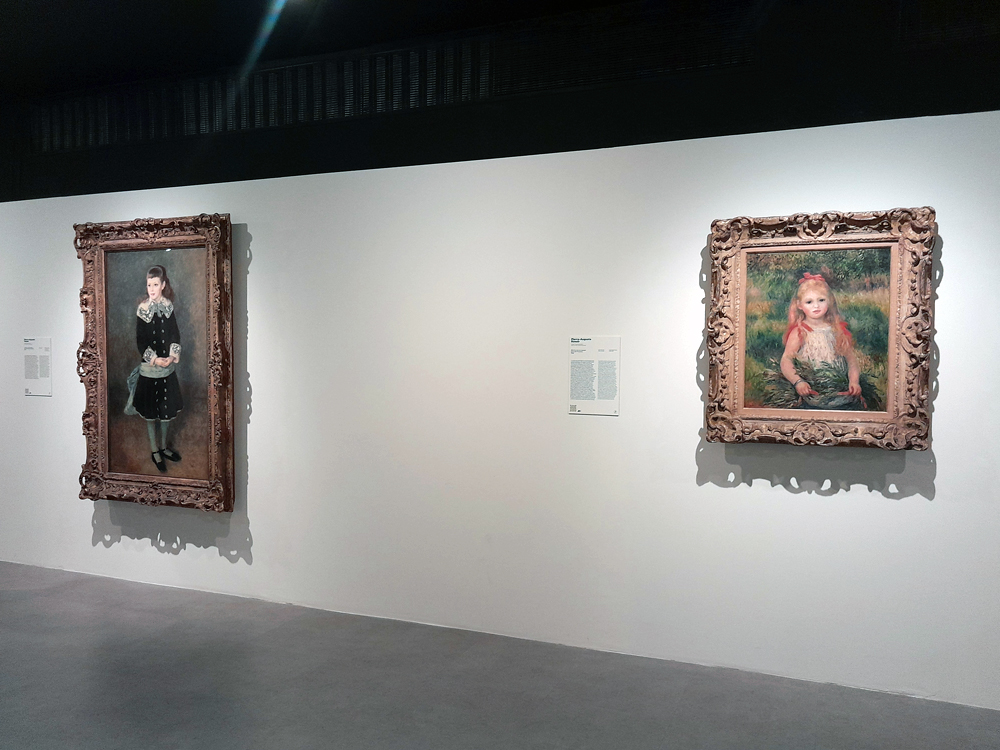Vista da exposição com dois retratos pintados por Renoir em molduras ornamentadas: à esquerda, figura em roupa escura com detalhes de renda; à direita, criança de cabelos claros em ambiente verde. Etiquetas informativas acompanham cada obra.