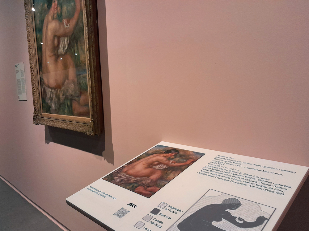 Pintura de figura nua sentada exibida em parede rosa, acompanhada de placa acessível com reprodução impressa, diagrama tátil e texto descritivo em português, permitindo diferentes formas de interação com a obra.