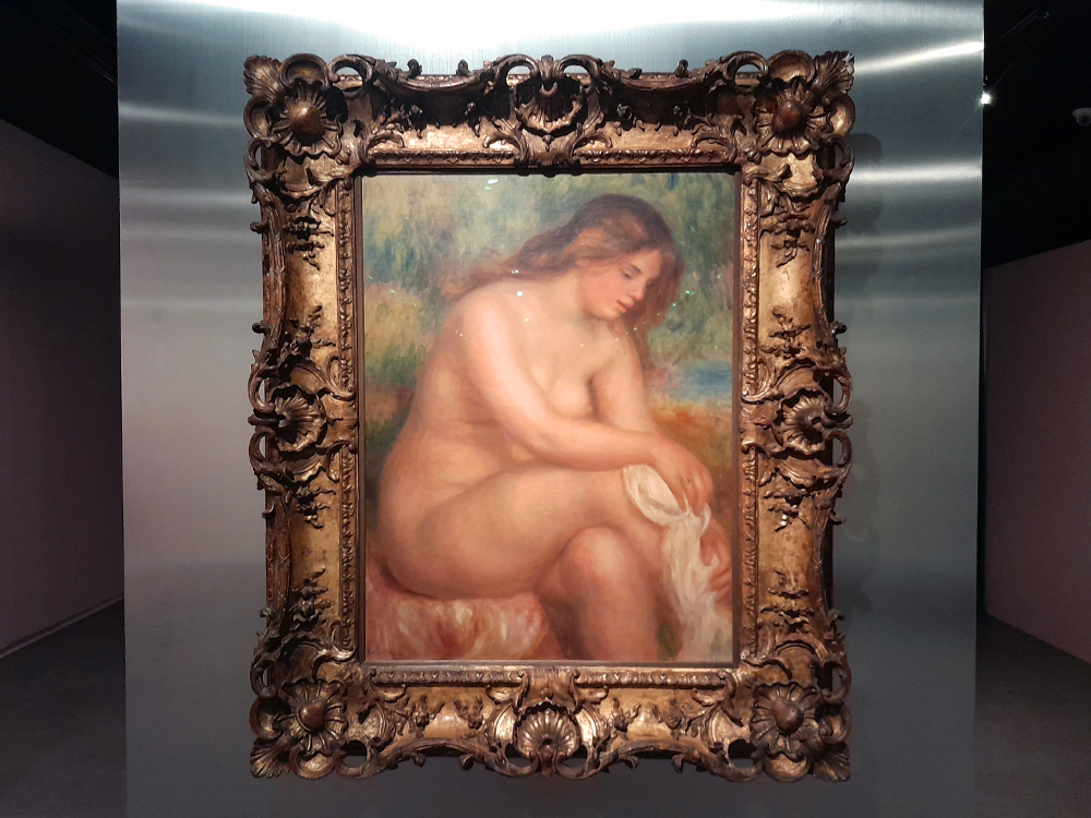 Pintura impressionista em moldura dourada mostrando figura nua sentada segurando pano branco, com fundo suave em tons de verde. A moldura apresenta entalhes florais detalhados que contrastam com as pinceladas fluidas da obra.