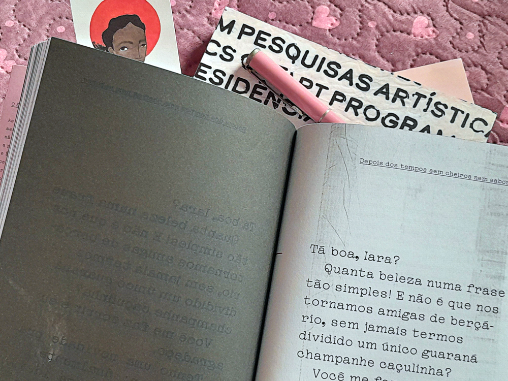 Livro Faca de Ponta, de Andrea Viviana Taubman e Érica Toledo, aberto sobre um tecido rosa texturizado com pequenos corações. A fotografia destaca uma página do romance com um trecho iniciado por Tá boa, Iara?, impresso em fonte que remete a máquina de escrever. Ao fundo aparecem um caderno com estampa tipográfica em preto e branco, uma caneta tinteiro rosa e um marcador de páginas ilustrado com o retrato estilizado de uma mulher.