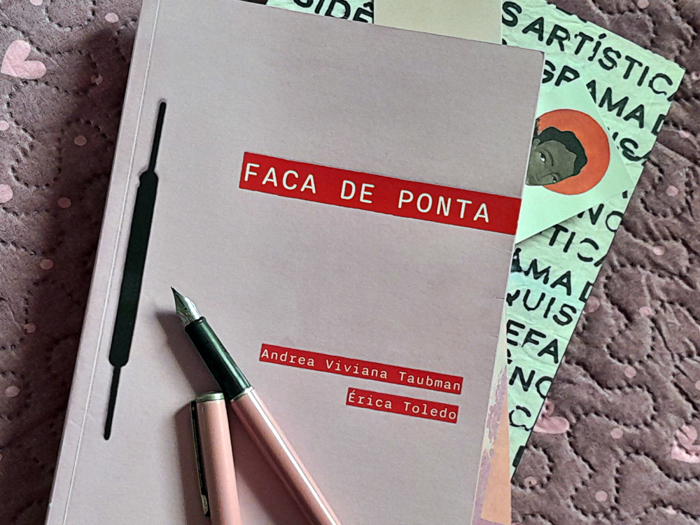 Exemplar do livro Faca de Ponta, de Andrea Viviana Taubman e Érica Toledo, fotografado de cima sobre um tecido roxo texturizado com pequenos corações. A capa rosa claro exibe o título em destaque sobre uma faixa vermelha e uma ilustração preta. Uma caneta tinteiro rosa repousa sobre o livro, enquanto ao fundo aparecem um caderno com estampa tipográfica e um marcador de páginas ilustrado.