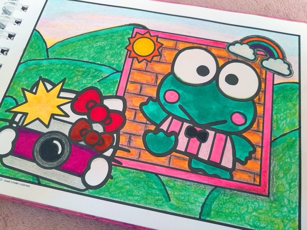 Página de livro de colorir da Hello Kitty mostrando Hello Kitty em um carrinho rosa e branco ao lado de Keroppi, que aparece em primeiro plano. Ao fundo há uma paisagem com colinas verdes, uma cerca de madeira laranja, sol amarelo e um arco-íris. A ilustração foi colorida com marcadores em tons vibrantes de rosa, verde, azul, laranja e amarelo, com textura visível do preenchimento manual.