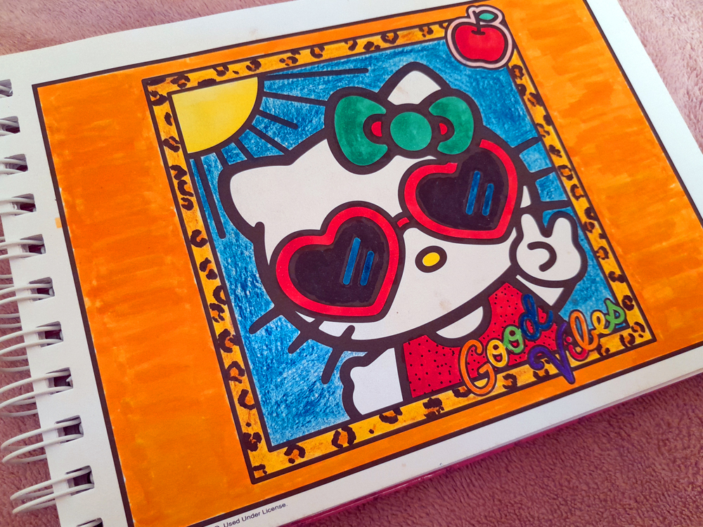 Página de livro de colorir da Hello Kitty mostrando a personagem usando grandes óculos escuros em formato de coração e fazendo um gesto de paz com uma das mãos. Hello Kitty veste uma roupa vermelha e está diante de um fundo azul, cercada por detalhes coloridos, com moldura laranja e pequenos elementos decorativos. A imagem foi preenchida com marcadores em tons intensos de azul, vermelho, laranja, verde e roxo.