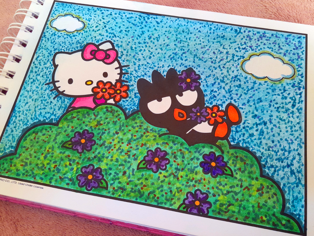 Página de livro de colorir da Hello Kitty mostrando Hello Kitty e Kuromi escondidas atrás de uma grande moita verde coberta por pequenas flores roxas. Hello Kitty segura flores vermelhas e laranjas, enquanto Kuromi aparece ao lado usando uma flor roxa na cabeça. O fundo é azul-claro com pequenas nuvens brancas, e toda a ilustração foi colorida com marcadores, criando um efeito pontilhado e texturizado especialmente no céu e na vegetação.