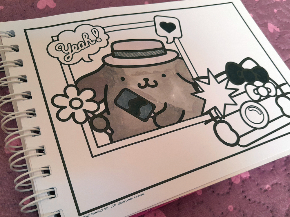 Página de livro de colorir mostrando Pompompurin usando um chapéu e segurando uma pequena flor, enquanto Hello Kitty aparece à direita fotografando a cena com uma câmera. Pompompurin está parcialmente colorido em tons de cinza, enquanto grande parte da página permanece sem colorir. A composição tem balões com as palavras Yeah! e um coração, além de uma explosão gráfica próxima à câmera.