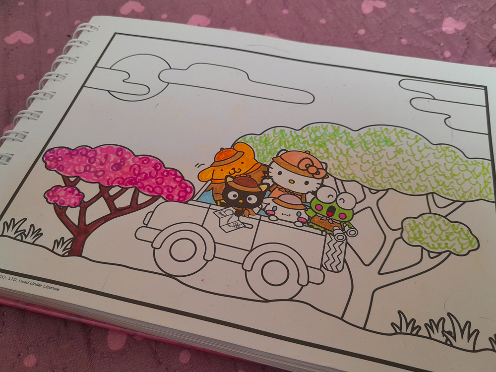 Página de livro de colorir mostrando Hello Kitty e seus amigos viajando em um pequeno carro conversível por uma paisagem arborizada. Hello Kitty, Kuromi, Pompompurin, Cinnamoroll e Keroppi aparecem sentados juntos no veículo, alguns usando chapéus e óculos. Apenas parte da ilustração está colorida: uma árvore à esquerda recebeu tons de rosa e marrom, as copas das árvores ao fundo foram preenchidas com verde-claro e os personagens aparecem parcialmente coloridos, enquanto o carro, a estrada e grande parte da paisagem permanecem sem preencher.