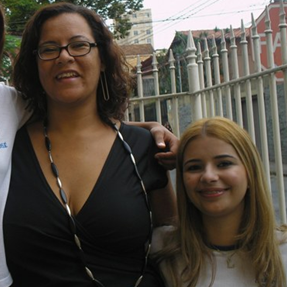 Fotografia antiga de Luly aos 17 anos, ainda adolescente, ao lado de sua professora de Química. Luly aparece à direita, com cabelos loiros compridos e usando uma camiseta clara, enquanto a professora está à esquerda, usando uma blusa preta e um colar comprido. As duas sorriem para a câmera em frente a um edifício, durante uma atividade ou visita escolar. Luly está posicionada em um degrau abaixo da professora.
