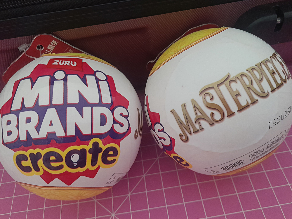Duas cápsulas colecionáveis Mini Brands sobre superfície rosa quadriculada: uma com rótulo colorido ZURU Mini Brands Create e outra branca com a palavra Masterpiece em dourado.