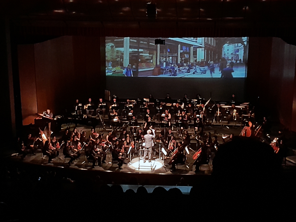 Palco com orquestra completa em apresentação em agosto de 2026, com músicos de cordas, sopros, metais e percussão acompanhados por maestro ao centro. Ao fundo, tela grande projeta cena de rua com pessoas caminhando e mesas externas, criando efeito de concerto multimídia.