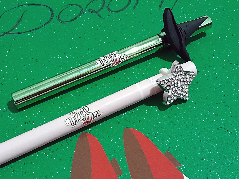 Dois delineadores temáticos coleção sobre fundo verde com glitter. O primeiro, vede, tem topo em formato de chapéu de bruxa, e o rosa claro traz uma estrela brilhante.