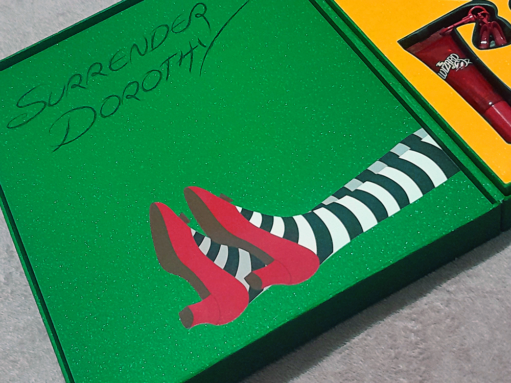 Caixa decorativa verde com glitter da coleção, trazendo ilustração das pernas da Bruxa do Leste com meias listradas e sapatos vermelhos, acompanhada da frase Surrender Dorothy escrita à mão, cenário das fotos de produtos. Ao lado, área amarela com tubo vermelho de gloss.