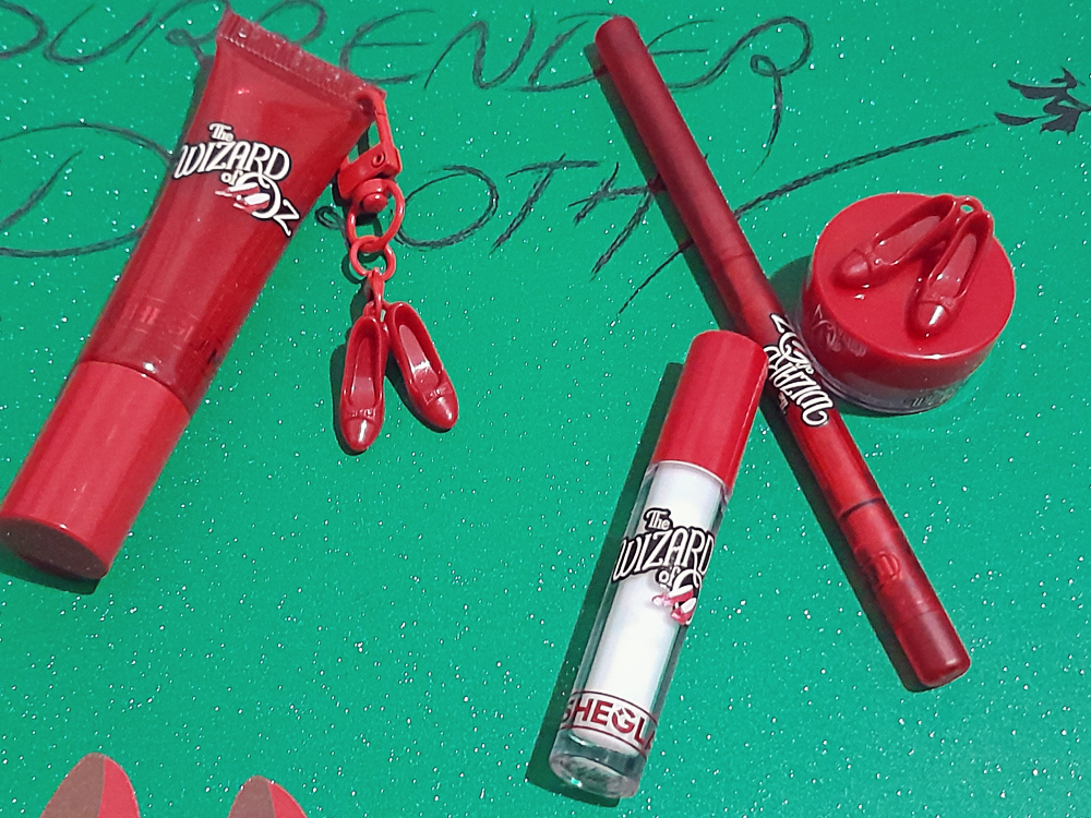 Conjunto de produtos labiais da coleção O Mágico de Oz da Sheglam sobre fundo verde com glitter. Inclui gloss vermelho com pingente de sapatinhos de rubi, lápis delineador labial vermelho, frasco pequeno com tampa vermelha e potinho redondo decorado com mini sapatinhos. Ao fundo, aparece a frase Surrender Dorothy escrita à mão.