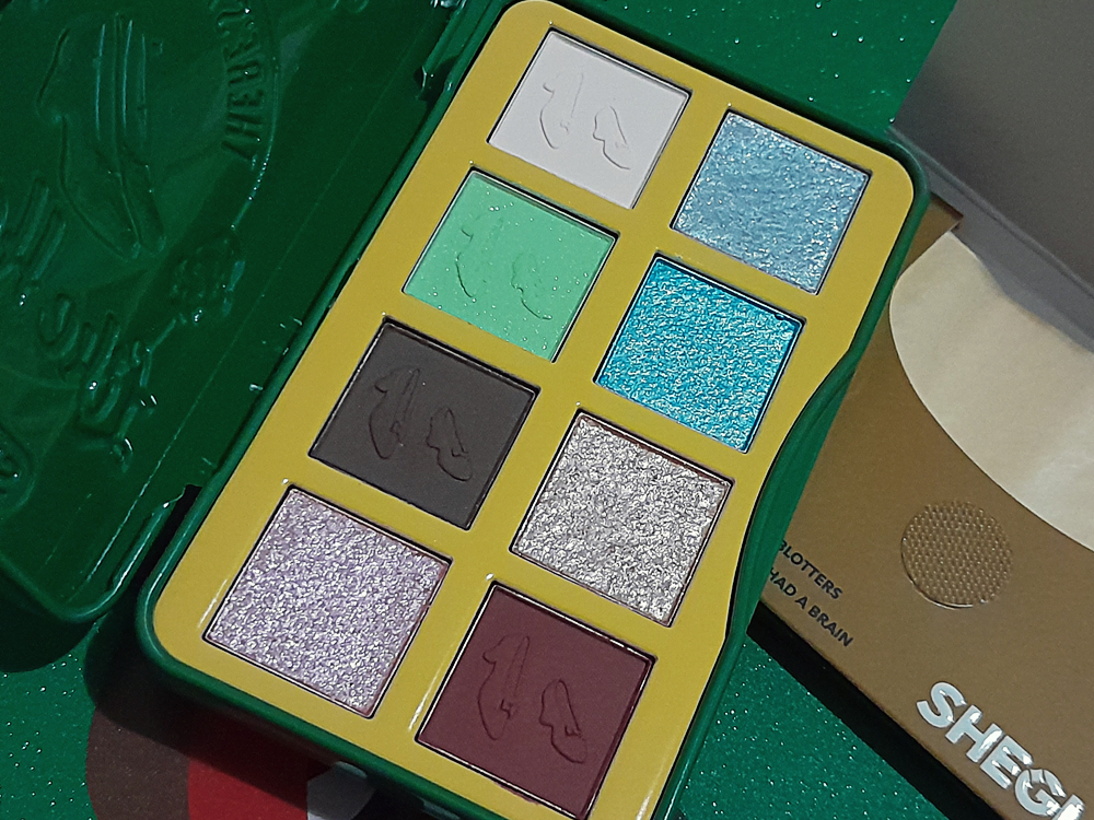 Paleta de sombras da Sheglam com oito cores em tons de verde, azul, branco, marrom, prata, vinho e rosa, organizada em grade duas grades de 4 cores dentro da lata verde que compõe a embalagem. Cada sombra tem relevo em formato de sapato, a paleta está ao lado do envelope de tiras anti-oleosidade aberto, onde se vê que elas têm tom creme..