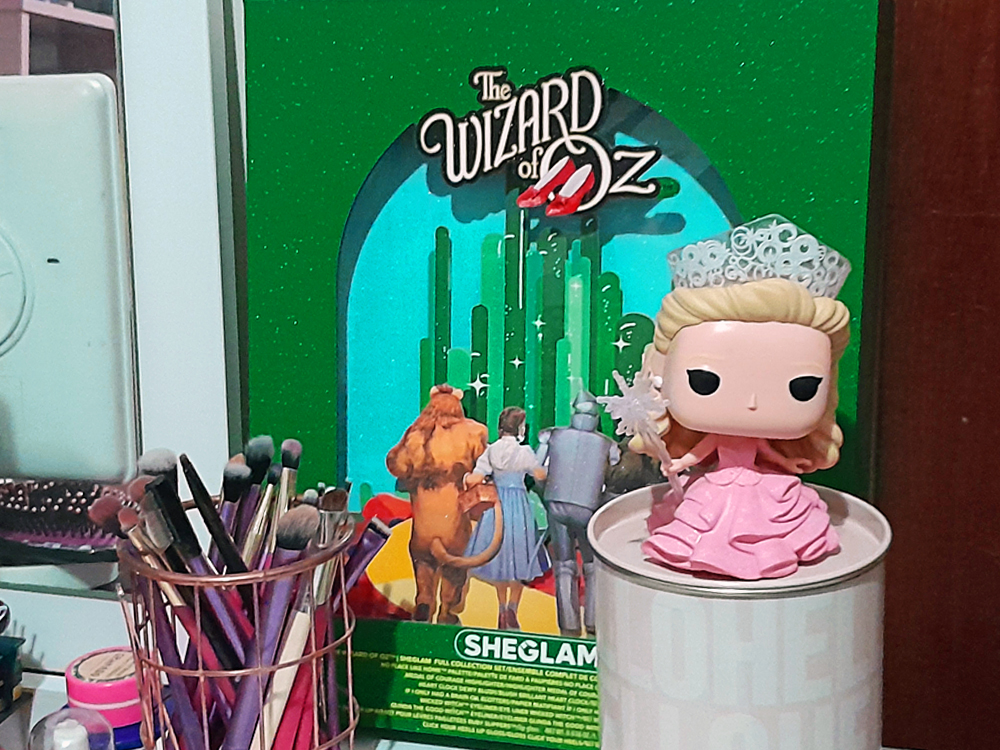 Display verde com glitter da coleção The Wizard of Oz da Sheglam mostrando Dorothy, o Leão, o Homem de Lata e o Espantalho caminhando rumo à Cidade das Esmeraldas. À frente, um Funko Pop da Glinda do filme Wicked com vestido rosa e varinha estrelada, ao lado de um pote com pincéis de maquiagem. A marca Sheglam aparece na base do display.