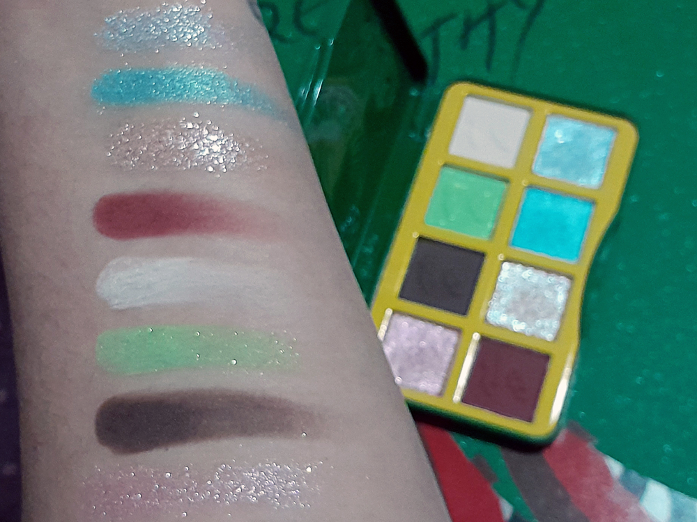 Braço com oito swatches da paleta de sombras mostrando tons de verde, azul, branco, marrom, rosa, vinho e prata. Ao lado, a paleta aberta revela as mesmas cores em formato retangular, com borda amarela e embalagem verde, posicionada sobre fundo verde com glitter.