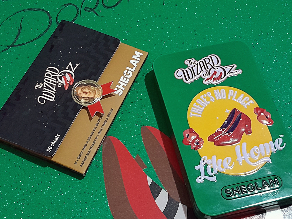 Kit de tiras anti oleosidade da inspirado no Espantalho de O Mágico de Oz, com embalagem preta e dourada exibindo o texto If Only I Had a Brain. Ao lado, uma lata verde da paleta de sombras decorada com elementos do filme, como os sapatos de rubi e a frase There’s No Place Like Home, ambos sobre fundo verde com glitter.