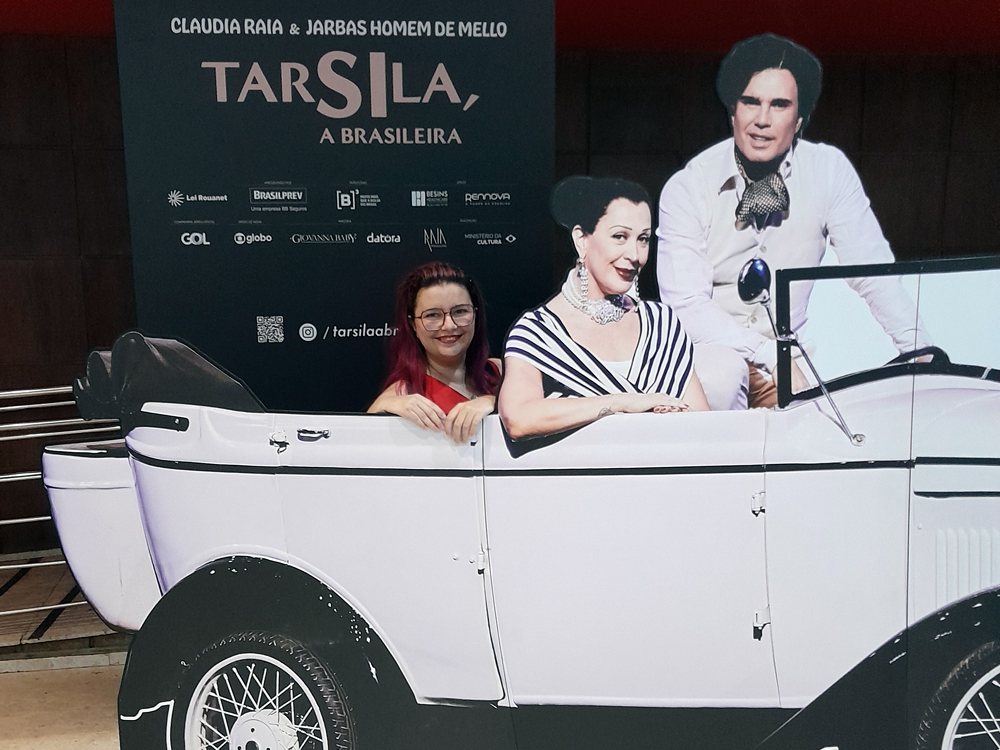 Uma foto minha posando atrás de um recorte em formato de carro conversível branco, ao lado de duas figuras impressas que simulam passageiros. Ao fundo, painel promocional da peça Tarsila, a Brasileira, com  logotipos de patrocinadores e o nome do espetáculo em destaque.