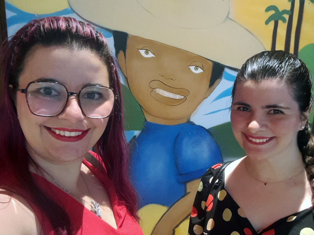 Duas mulheres, Luly e Daninha, sorrindo em frente a um mural colorido com personagem de chapéu grande, camisa azul e fundo com palmeiras e céu vibrante. Uma delas usa roupa vermelha e óculos; a outra veste roupa preta com estampa de bolinhas coloridas.
