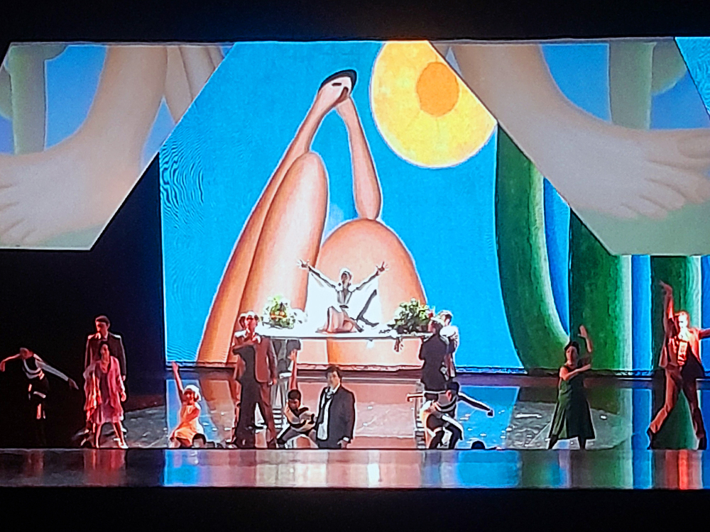 Cena de Tarsila, a Brasileira com elenco distribuído pelo palco em poses expressivas diante de projeção colorida inspirada na tela Abaporu, com figuras alongadas, sol amarelo e elementos verdes. A personagem central está elevada em plataforma cercada por plantas.