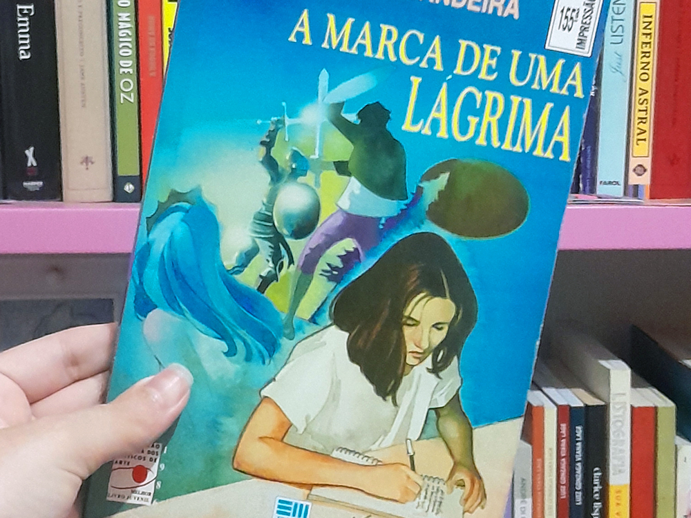 Fotografia do livro A Marca de Uma Lágrima sendo segurado por uma mão diante de uma estante de livros. A capa, em tons de azul, mostra uma jovem escrevendo em um caderno, além de figuras e silhuetas que remetem à narrativa. Ao fundo, aparecem livros organizados em prateleiras.