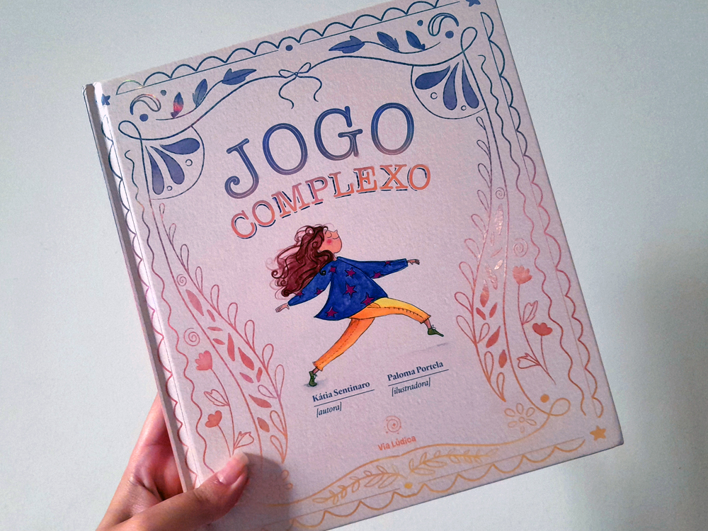 Fotografia do livro Jogo Complexo, de Kátia Sentinaro com ilustrações de Paloma Portela, sendo segurado por uma mão diante de uma parede clara. A capa em tons suaves de rosa, azul e dourado apresenta uma moldura decorativa com elementos florais e uma personagem ilustrada no centro, em movimento, usando roupas coloridas.