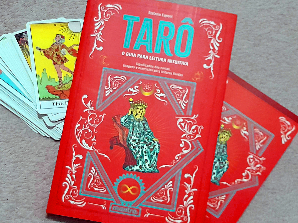 Fotografia do livro Tarô: O Guia para Leitura Intuitiva, de Stefanie Caponi, apoiado sobre uma superfície clara. A capa vermelha traz detalhes ornamentais em branco e azul-turquesa e a ilustração de uma figura sentada em um trono. Ao lado do livro, há um baralho de tarô parcialmente aberto, com uma carta THE FOOL visível no topo.