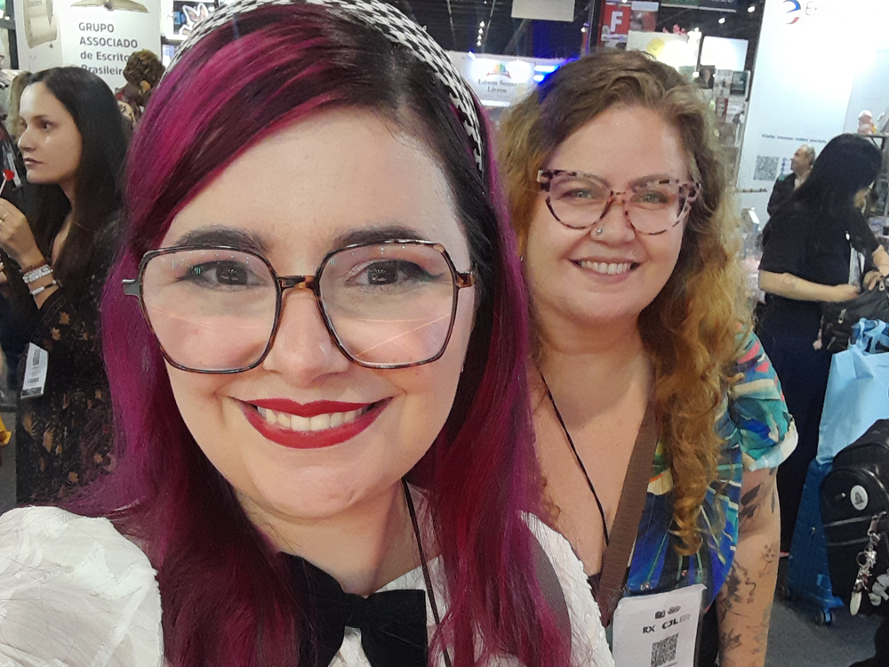 Duas mulheres posam sorrindo. Luly Lage tem cabelos rosa-choque, usa óculos, blusa branca com laços preto, tiara preto e branca e batom vermelho. Mari Mahé tem cabelos loiros cacheados, óculos, blusa estampada colorida. Ao fundo, estandes e visitantes do evento.