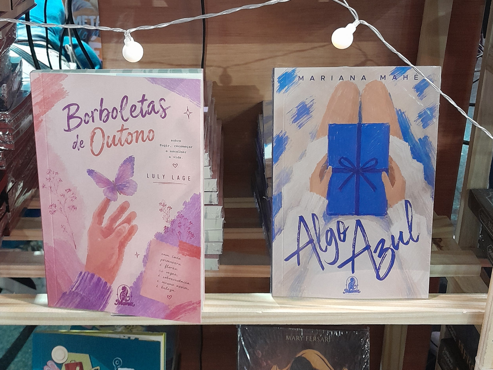 Dois livros expostos em prateleira com luzes. O primeiro é Borboletas de Outono, capa rosa com ilustração de mão estendida e borboleta, título em branco e subtítulo sobre fugir, recomeçar e escolher a vida. O segundo é Algo Azul, capa em tons de azul e bege com figura segurando uma caixa azul com laço, transmitindo clima delicado e romântico.