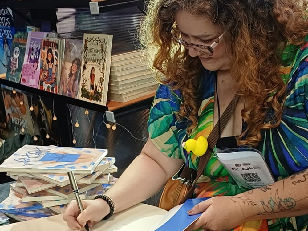 Mari Mahé assina livros em mesa de evento. À frente, pilhas de livros ilustrados; atrás, estante com títulos diversos.