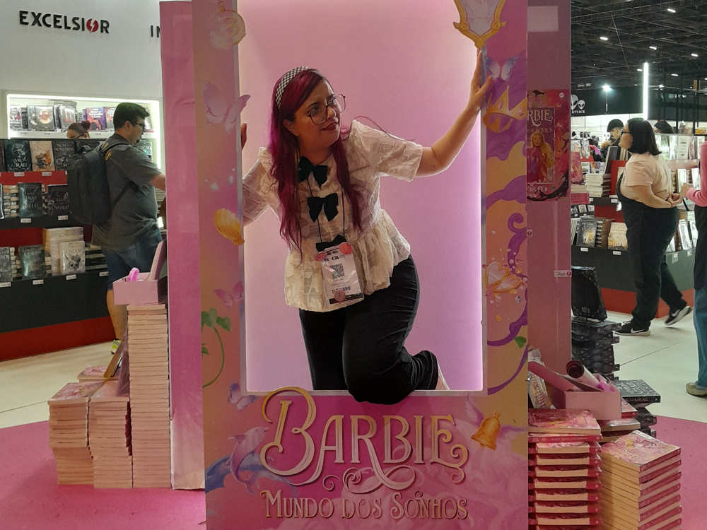 Luly posando dentro de estrutura rosa decorada como caixa da boneca Barbie, com borboletas e brilhos. Ao fundo, estantes de livros e visitantes da Bienal de São Paulo.