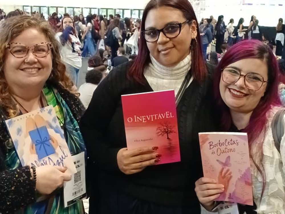 Três autora posam segurando seus livros: Algo Azul, O Inevitável e Borboletas de Outono. Ambiente movimentado de feira literária com banner azul ao fundo exibindo gêneros como fantasia, romance e suspense.