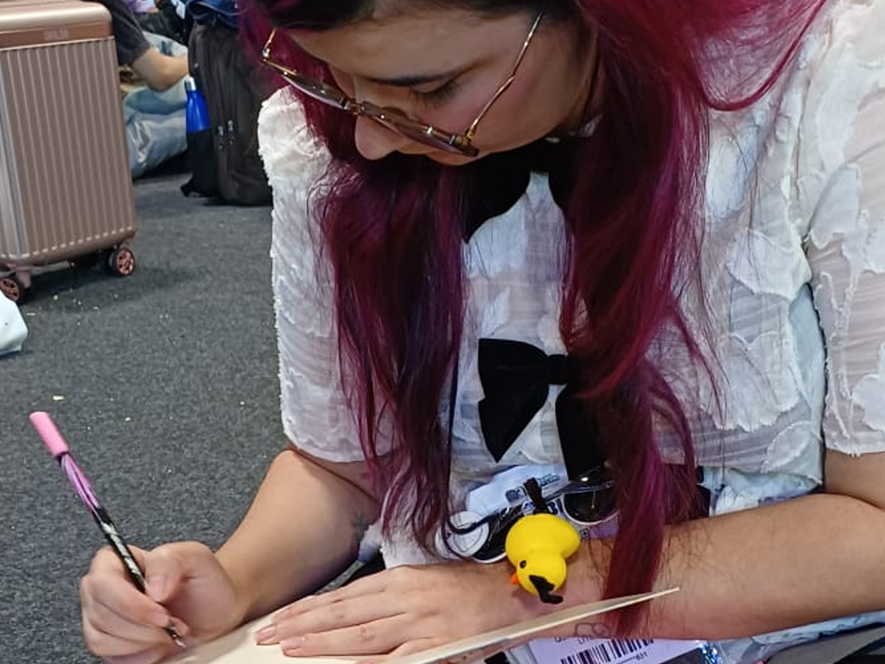Luly sentada no chão da Bienal de São Paulo autografando um livro com caneta rosa. Ao fundo, malas e visitantes descansando no corredor.