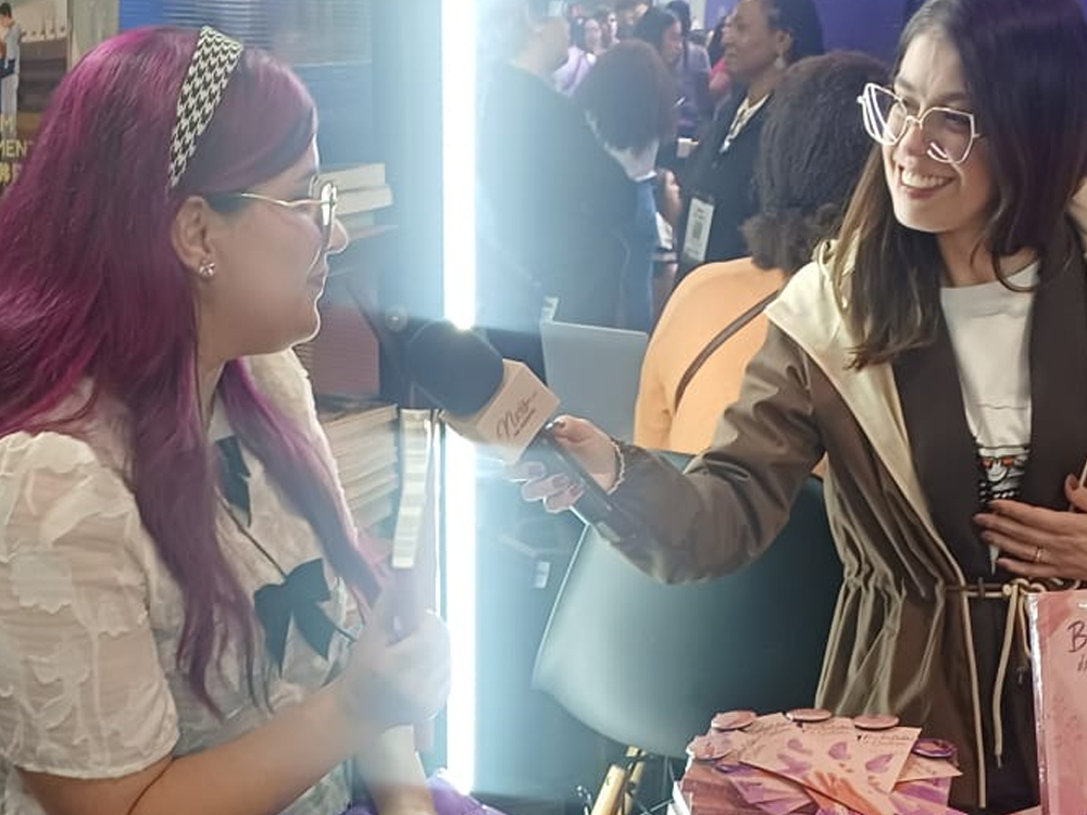 Luly segura livro enquanto conversa com entrevistadora de óculos e jaqueta marrom, que segura. Mesa com livros e visitantes ao fundo reforça clima de cobertura jornalística da Bienal.