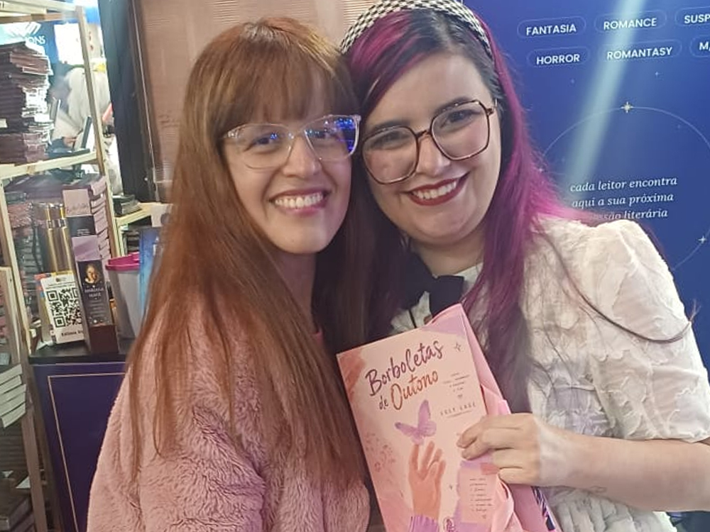 Leitora posando ao lado de Luly sorrindo, ambas segurando um exemplar de Borboletas de Outono. Ao fundo, estande com livros e banner azul com gêneros literários e frase: cada leitor encontra aqui a sua próxima paixão literária.