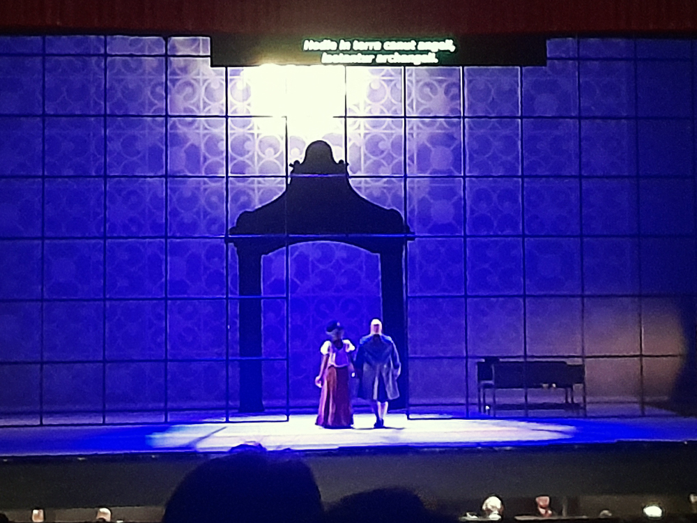 Dois intérpretes, uma mulher e um homem, em trajes de época atuam de costas, de braços dados, sob grande arco cenográfico, diante de painel quadriculado iluminado em azul e roxo. A luz cria atmosfera solene enquanto o texto projetado acima funciona como legenda da ópera. À direita, mesa de madeira compõe o ambiente dramático.